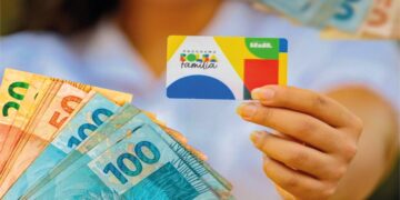 Cartão do Bolsa Família sendo segurado por uma mão, com notas de dinheiro ao fundo