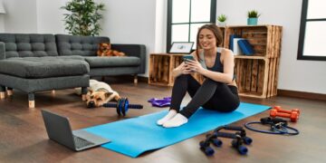 Mulher praticando exercícios em casa com smartphone, tapete de yoga e acessórios de ginástica